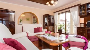 A living room with a sofa set, a glass-top coffee table, and a large mirror. There is a view of the outside through a window with curtains. - Holiday apartment with balcony and sea view (Klimno)