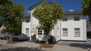 Ein weißes Gebäude mit Baum davor, ein Auto auf der Straße und ein klarer, blauer Himmel.