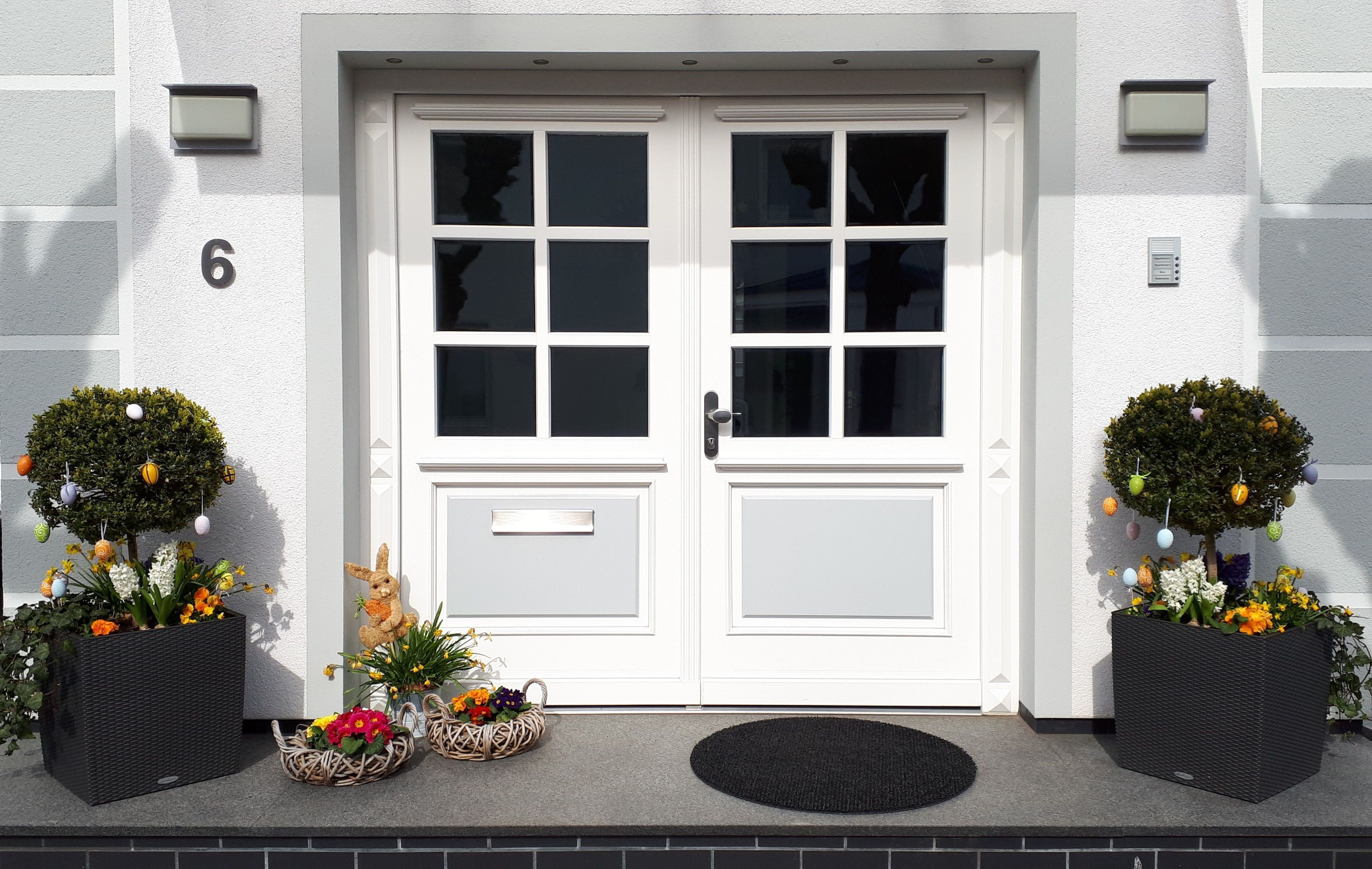 A white double door with a number '6' above it, two potted plants with flowers and ornaments, and a black mat at the entrance.