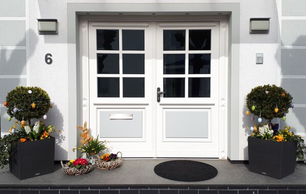 A white double door with a number '6' above it, two potted plants with flowers and ornaments, and a black mat at the entrance.