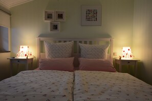 A double bed with floral bedding, two bedside lamps, a white headboard, and framed pictures on the wall.
