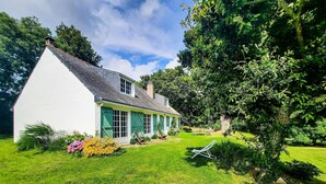 Una casa blanca con contraventanas verdes, techo inclinado y jardín con césped y flores.