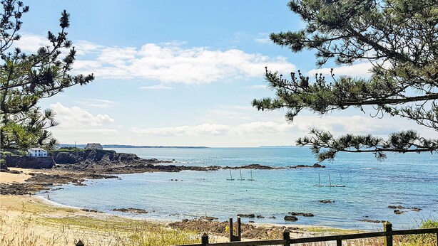 Vista costera con valla de madera, playa arenosa, costa rocosa y una casa al fondo.