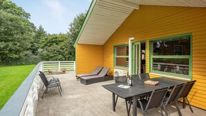 En gul bygning med en overdækket terrasse, hvor der er et bord, stole, en ventilator og et vindue med grøn ramme.