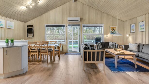 En rummelig stue med højt til loftet, trævægge, et spiseområde med bord og stole, en sofa, og et køkkenområde med en bordplade og skabe.