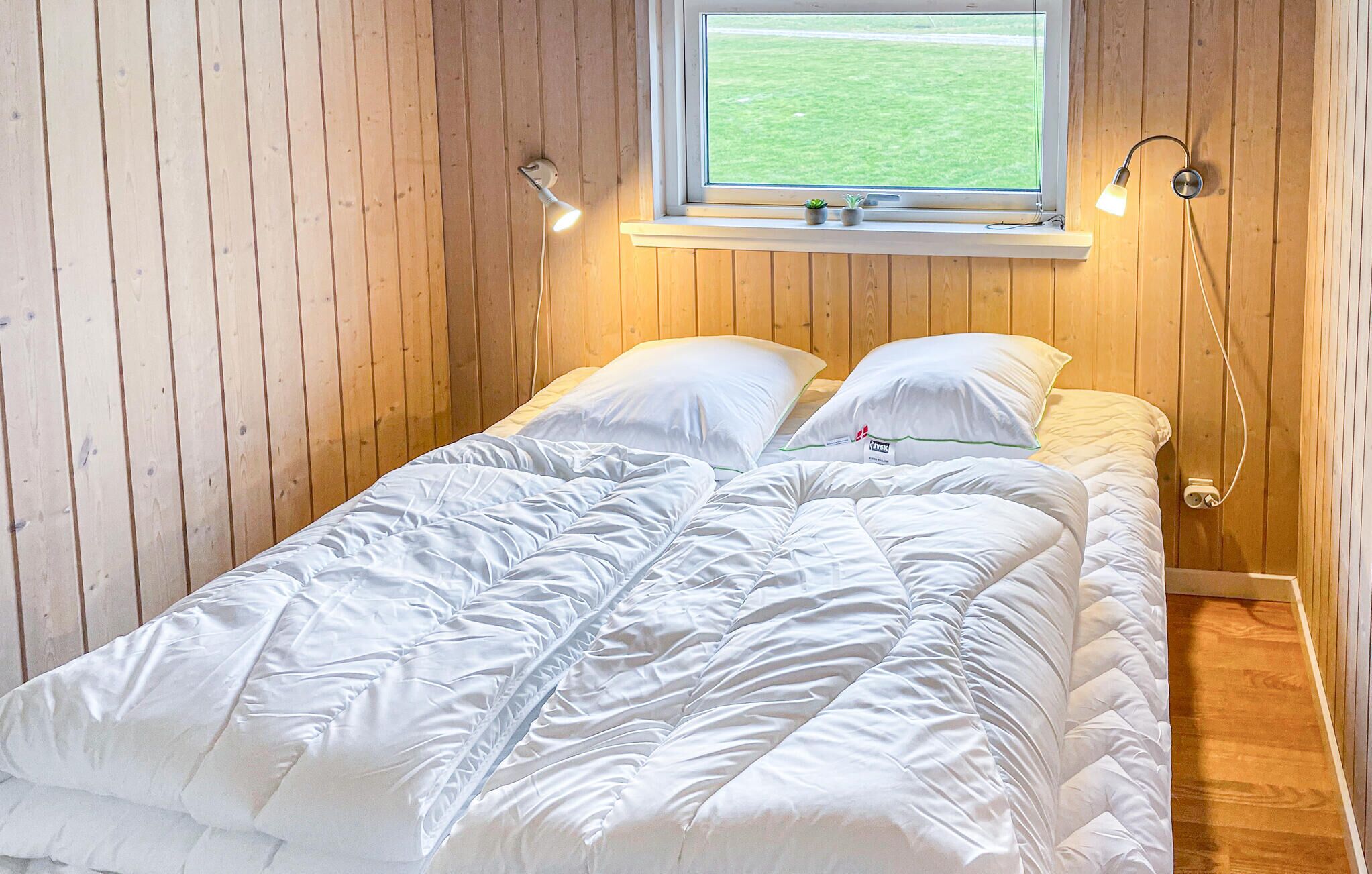 A single bed with a white duvet and pillows in a room with wooden walls and a window with a view of a grassy field.