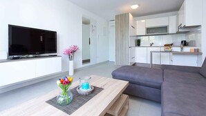 A modern living room with a flat-screen TV, a grey sofa, a wooden coffee table, and a kitchen area with white cabinets and a microwave.
