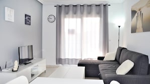 Una sala moderna con un sofá gris, una mesa de centro blanca, un televisor sobre un mueble blanco y un amplio ventanal con cortinas grises.