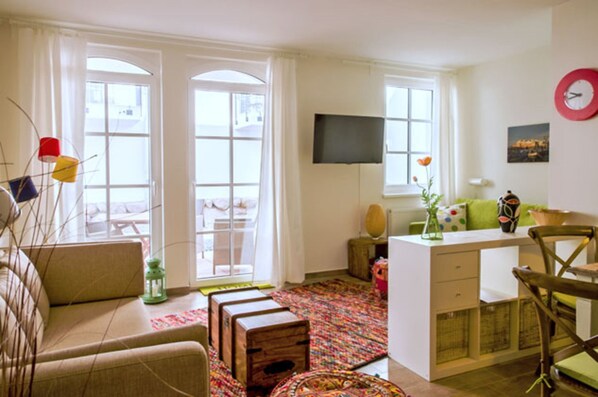 Un salon chaleureux avec un canapé, une petite table et un téléviseur. De grandes fenêtres sont ornées de rideaux, et la pièce est agrémentée d’un tapis à motifs et d’une armoire en bois.