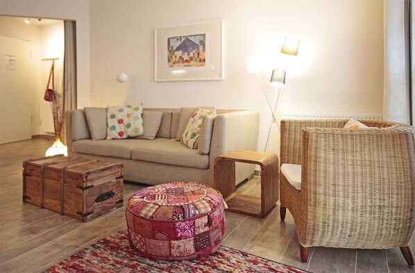 A living room with a sofa, a wicker chair, a wooden chest, and a round ottoman.
