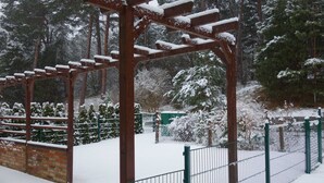 A snow-covered pergola structure in a park with a brick wall and a metal fence.