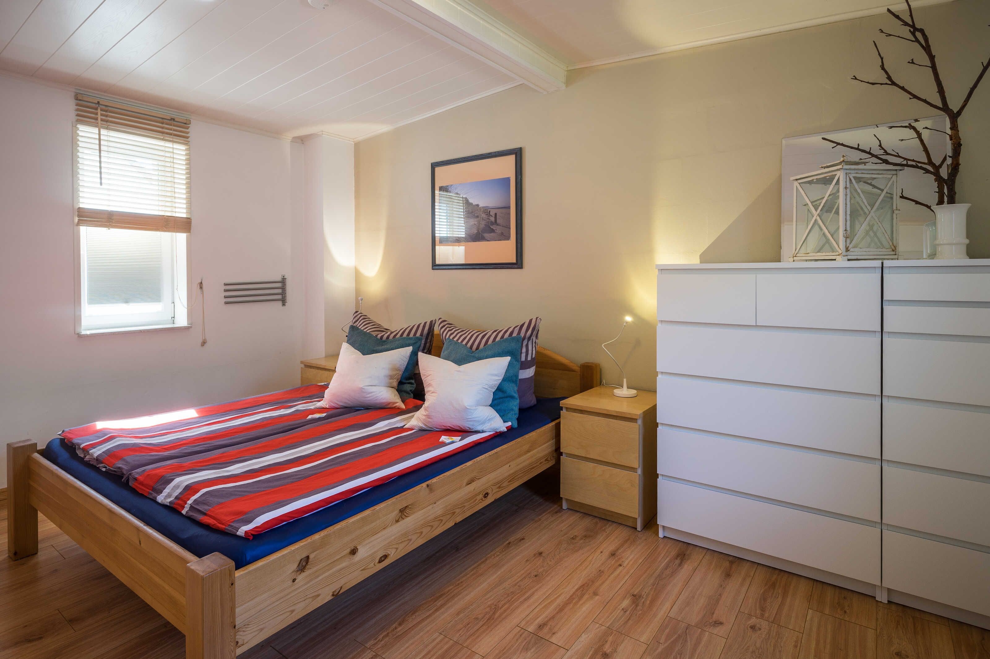 Quarto com cama de madeira, mesa de cabeceira, cômoda branca, quadro na parede e janela com persianas.