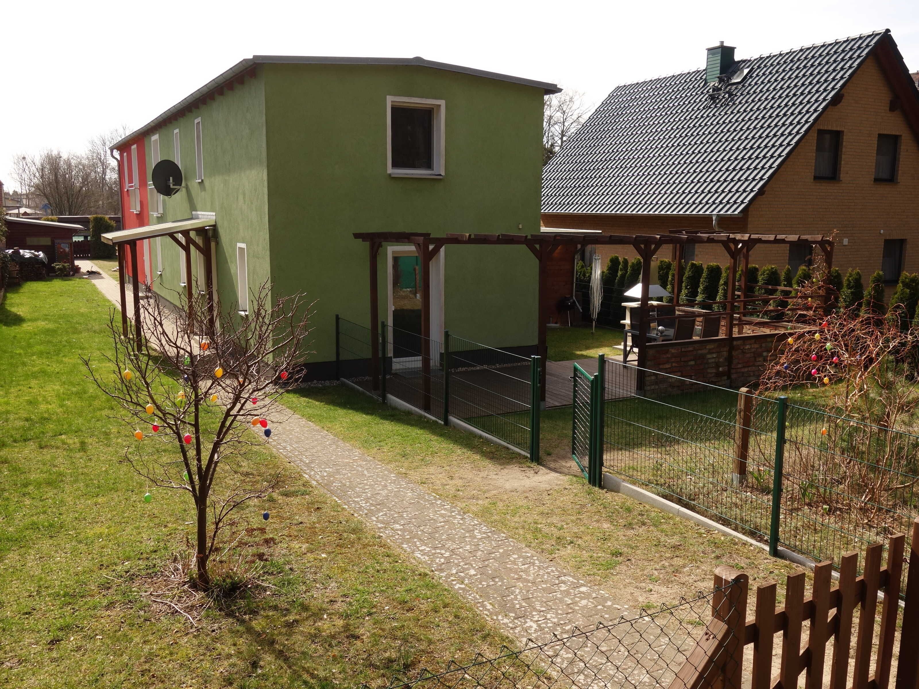 Ein grünes Gebäude mit einem kleinen Fenster und einer Tür, einem gepflasterten Weg und einem eingezäunten Bereich mit einer Pergola.