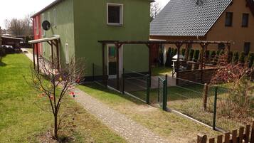 Ein grünes Gebäude mit einem kleinen Fenster und einer Tür, einem gepflasterten Weg und einem eingezäunten Bereich mit einer Pergola.