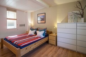 Une chambre à coucher avec un lit en bois, une table de chevet, une commode blanche, un tableau encadré au mur et une fenêtre avec des stores.