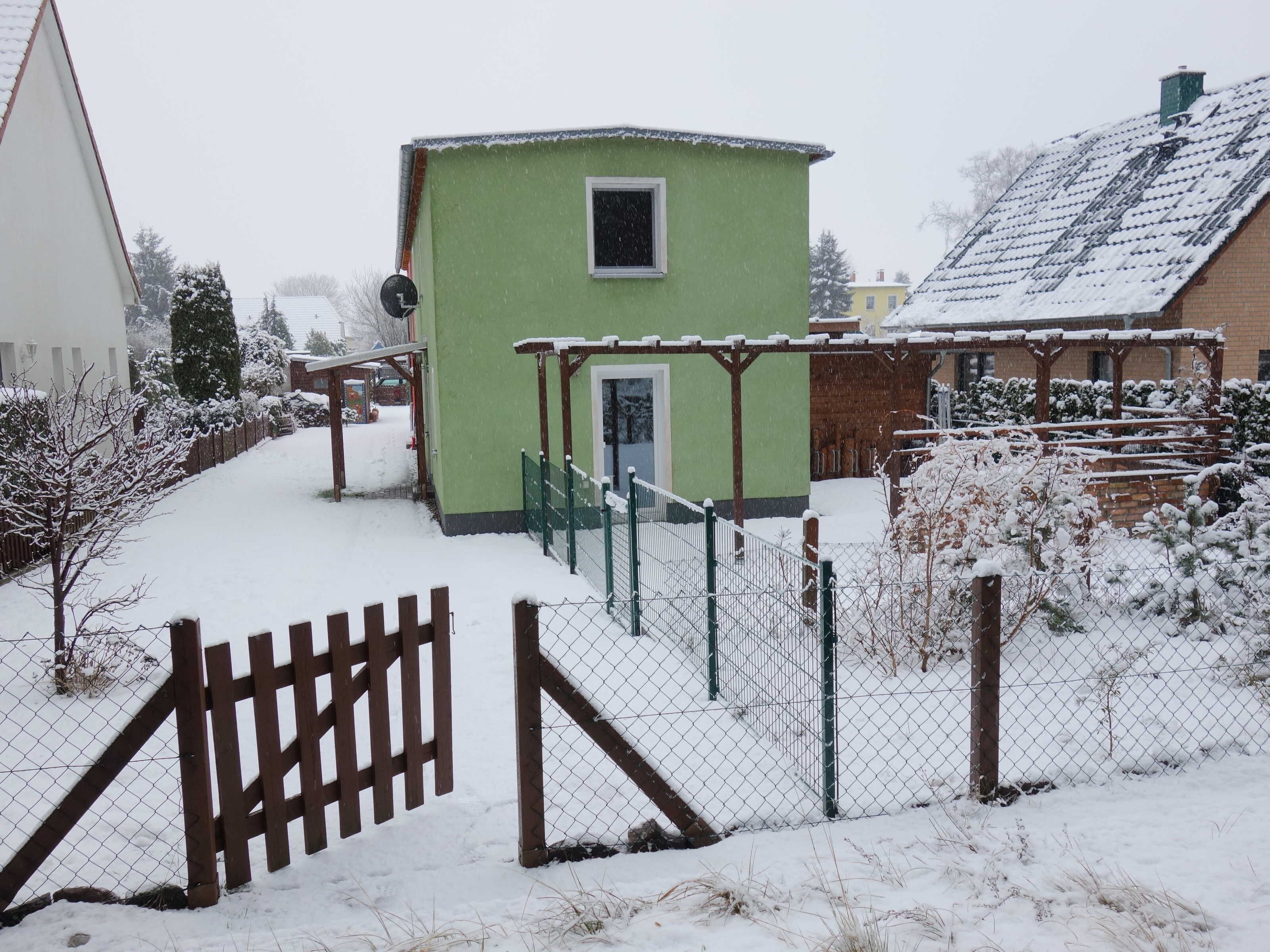 Una casa verde con una ventana pequeña, un área cercada y alrededores cubiertos de nieve.