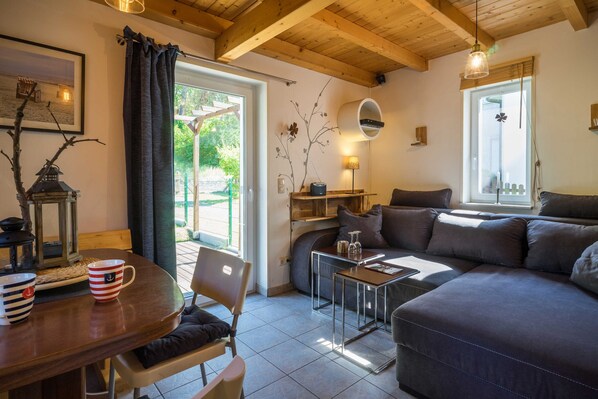 A cozy living room with a sofa, a dining table, and a coffee table. There is a window with a view of greenery outside.