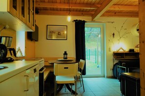 Une cuisine avec une table à manger, des chaises, un luminaire suspendu et une vue sur l’extérieur, visible à travers une porte.