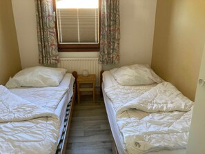 A small, single-room hotel room with two single beds, a wooden nightstand, a radiator, and a window with curtains.