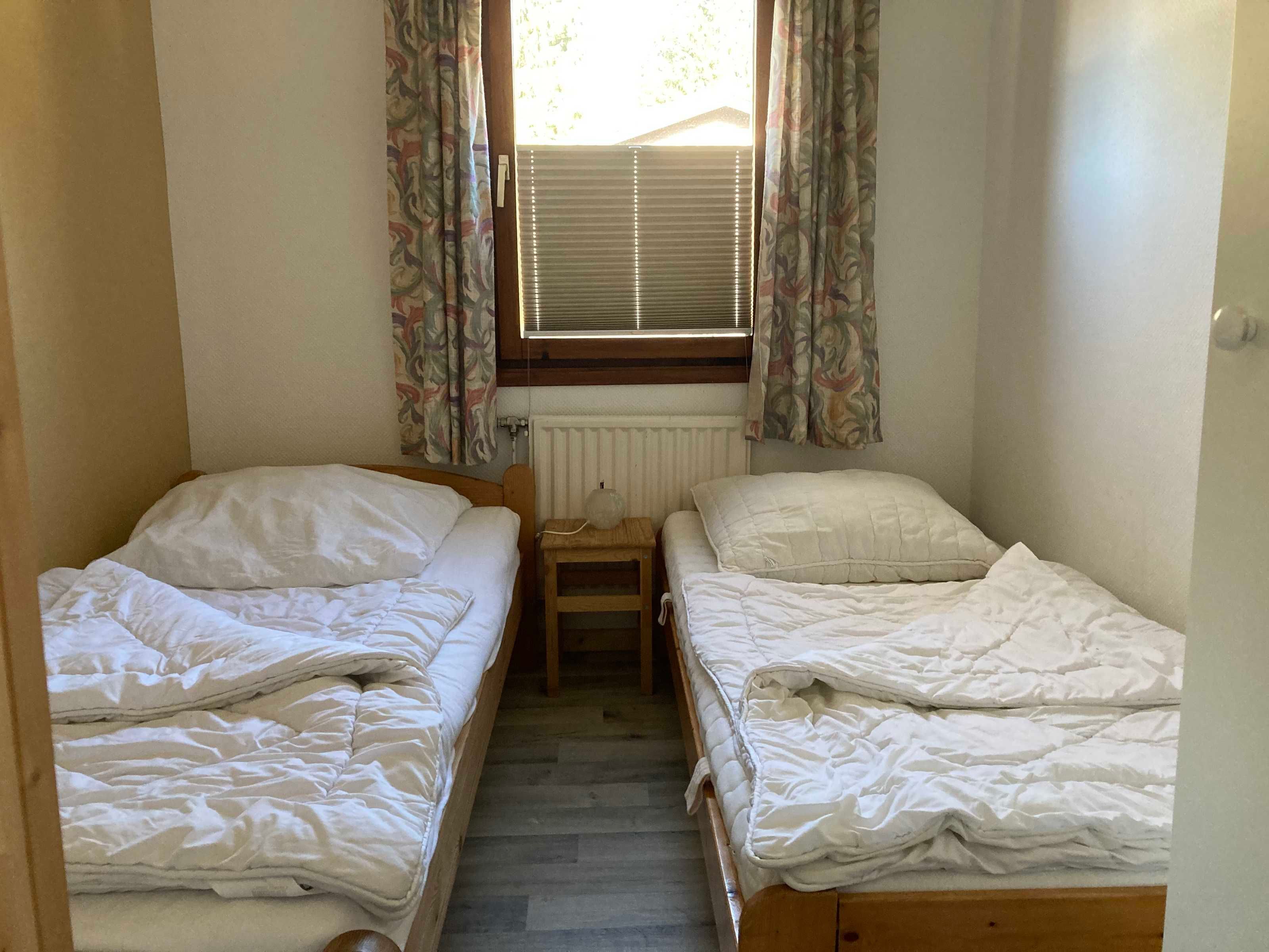 Una habitación pequeña con dos camas individuales, una mesita de noche de madera, un radiador y una ventana con cortinas.