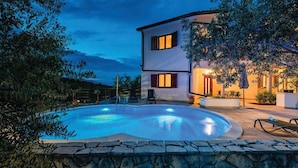 Una casa de dos pisos con piscina, pared de piedra y mobiliario exterior.