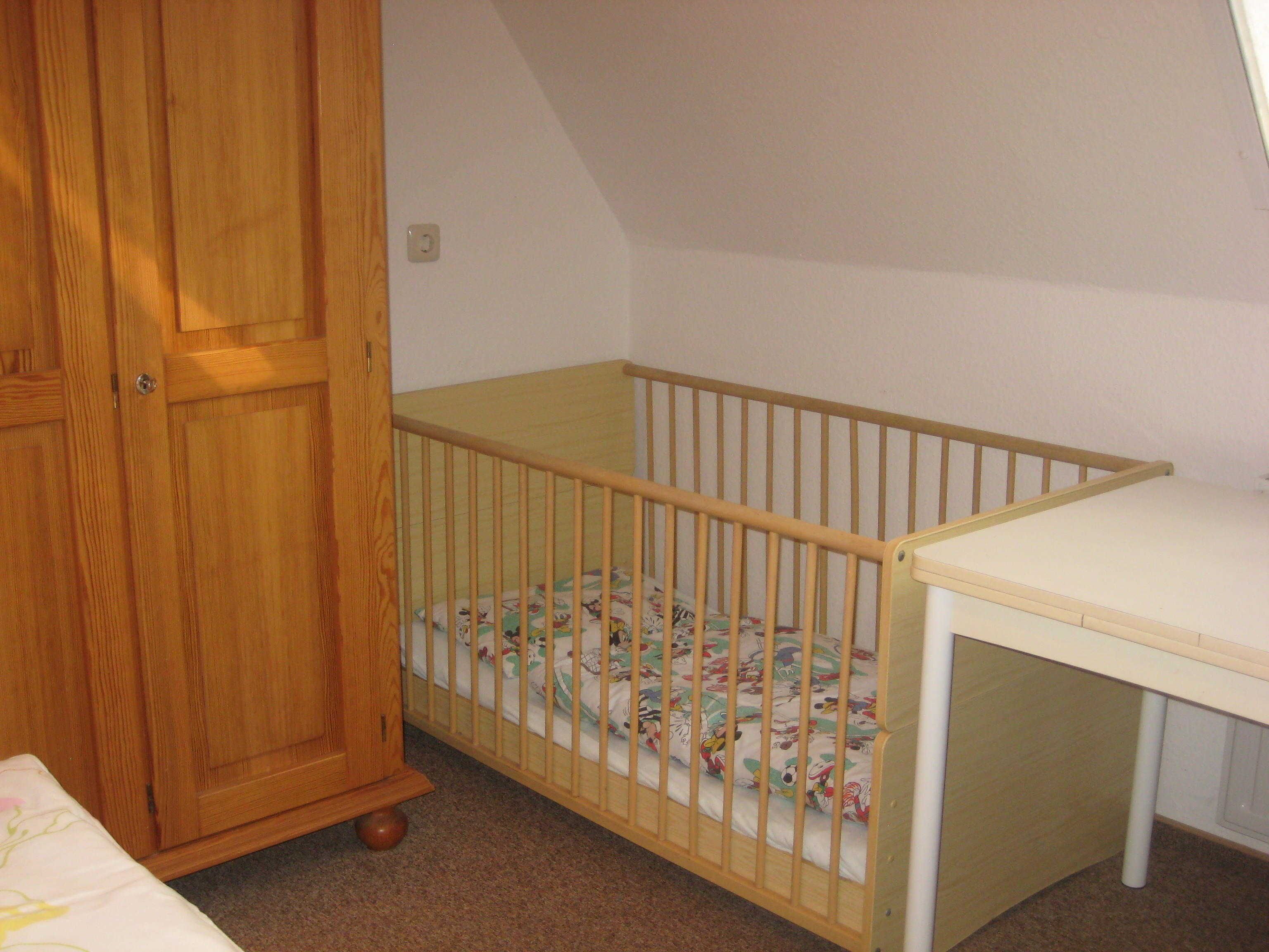 Una habitación con un armario de madera, una cuna con colchón floral y una mesita pequeña.