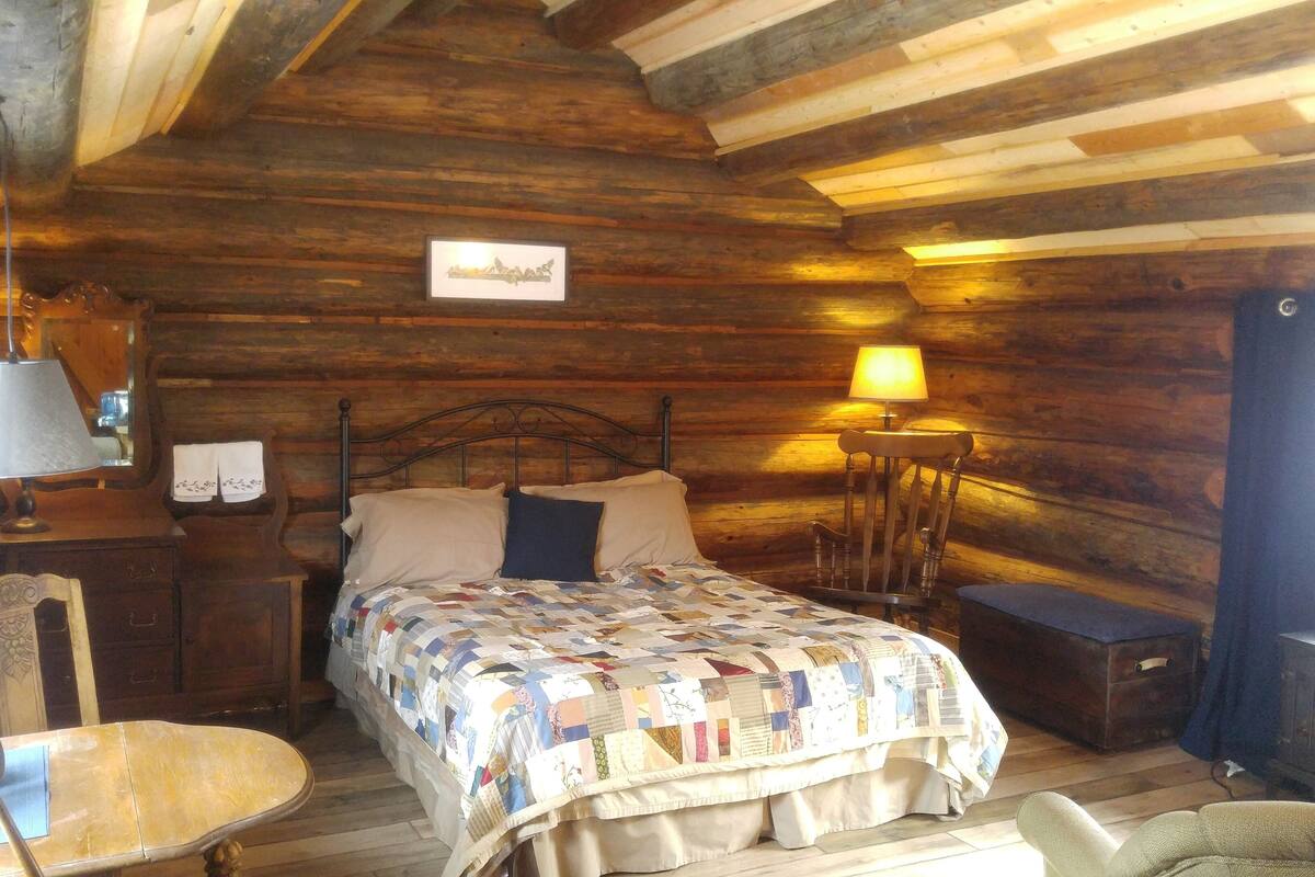 Ett mysigt sovrum i en stuga med träpanel, inrett med en säng, en stol, ett bord, en lampa och en öppen spis.