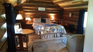 A cozy log cabin bedroom with a bed, two bedside tables with lamps, a wooden chair, and a small round table. - Wolfwood Guest Ranch (Clearwater)
