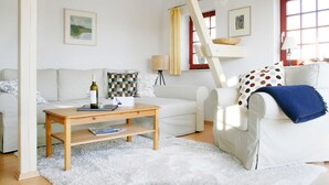 A living room with a white sofa, a wooden coffee table, and a white rug. There is a window with a red frame and a lamp on a side table. - Remise Whg. 13 - Villa Bellevue (Heringsdorf)