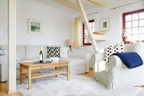 A living room with a white sofa, a wooden coffee table, and a white rug. There is a window with a red frame and a lamp on a side table.