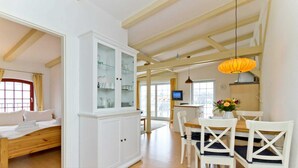 A spacious room with a wooden bed, a white cabinet with glass doors, a dining area with a table and chairs, and a kitchen in the background. - Remise Whg. 13 - Villa Bellevue (Heringsdorf)
