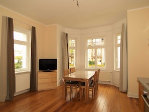 Un comedor con mesa y sillas de madera, televisor sobre un mueble y ventanas grandes con cortinas.