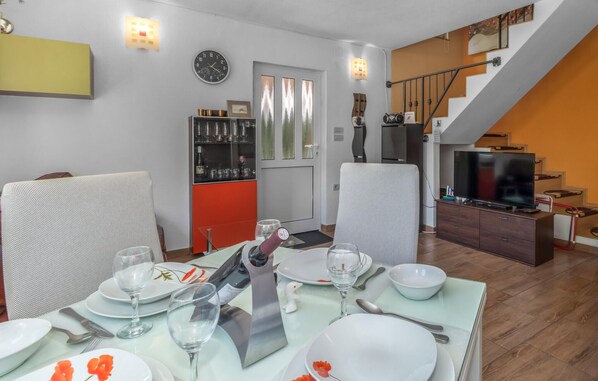 A dining area with a table set for four, a bar area with wine glasses, a clock on the wall, and a staircase leading to a living area with a TV.