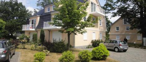 A residential building with a balcony, a driveway, and a lawn with bushes.