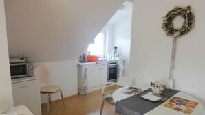 Een kleine keuken met een magnetron, oven en spoelbak, een eethoek met een tafel en stoelen, en een klein houten tafeltje met keukengerei.