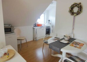A compact kitchen with a microwave, oven, and sink, a dining area with a table and chairs, and a small wooden table with kitchen utensils. - Haus Bernstein 5 - Apartment - Apartment Bernstein 5 (Graal-Müritz)