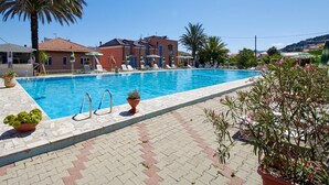 En swimmingpool med liggestole og parasoller, omgivet af plantekasser og en bygning med et tagtækket tag.