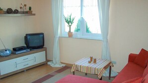 Una sala de estar con un sofá, una televisión, una mesita, una ventana con una planta y una estantería con objetos decorativos.