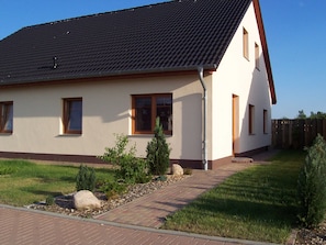 Une maison de plain-pied avec un toit en tuiles, un chemin de briques menant à l’entrée et un petit jardin agrémenté d’arbustes et d’un rocher.