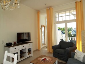 Un salon avec un téléviseur, des meubles blancs, une table basse en bois et une vue dégagée sur l’extérieur grâce à de grandes fenêtres.