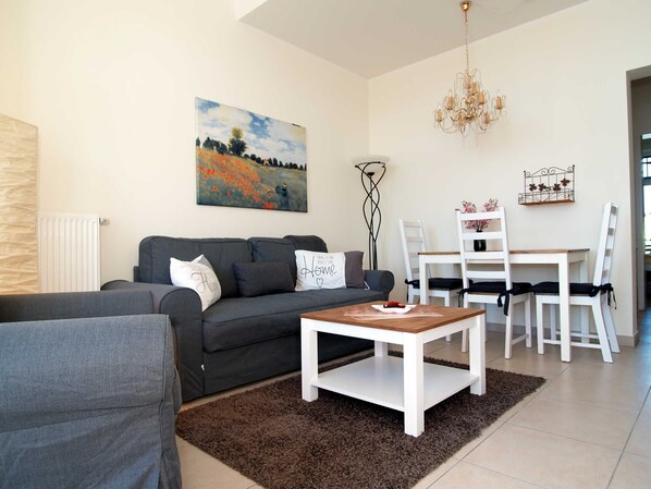 A modern living room with a grey sofa, a coffee table, a dining table with chairs, a chandelier, and a painting on the wall.