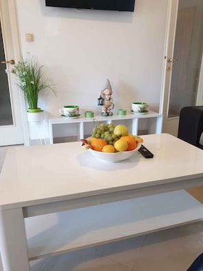 Et hvidt bord med en skål frugt og en fjernbetjening. En lille figur og et grønt lys står på en hvid hylde bag bordet.