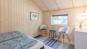 A room with wooden walls, a bed, two chairs, a small table, a window with a view, and a framed picture on the wall. - Stunning home in Nexø with kitchen (Nexø)