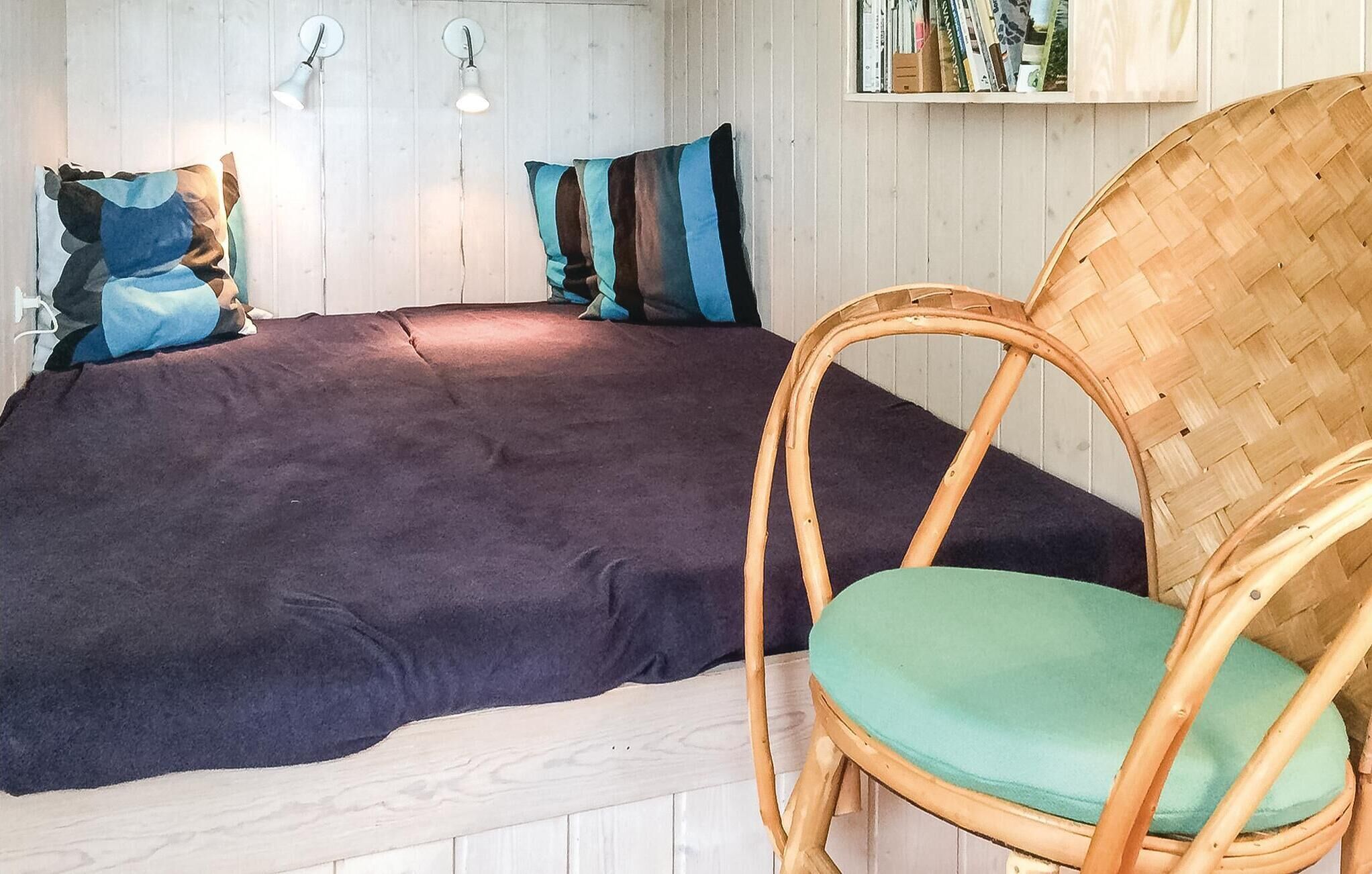 A bed with a dark cover, a wicker chair with a green cushion, and two wall-mounted lamps.