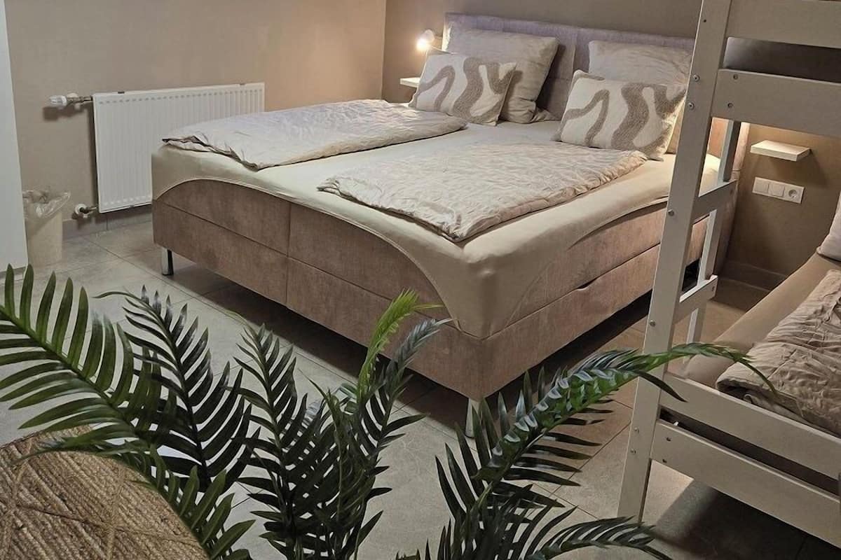 Un dormitorio con una cama doble, una ventana con persianas, un radiador, una mesita de noche con lámpara y una cama litera.