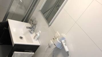 Ein modernes Badezimmer mit Duschkabine aus Glas, einem weißen Waschbecken inklusive Spiegel, einem Handtuchhalter und einem an der Wand befestigten Föhn.