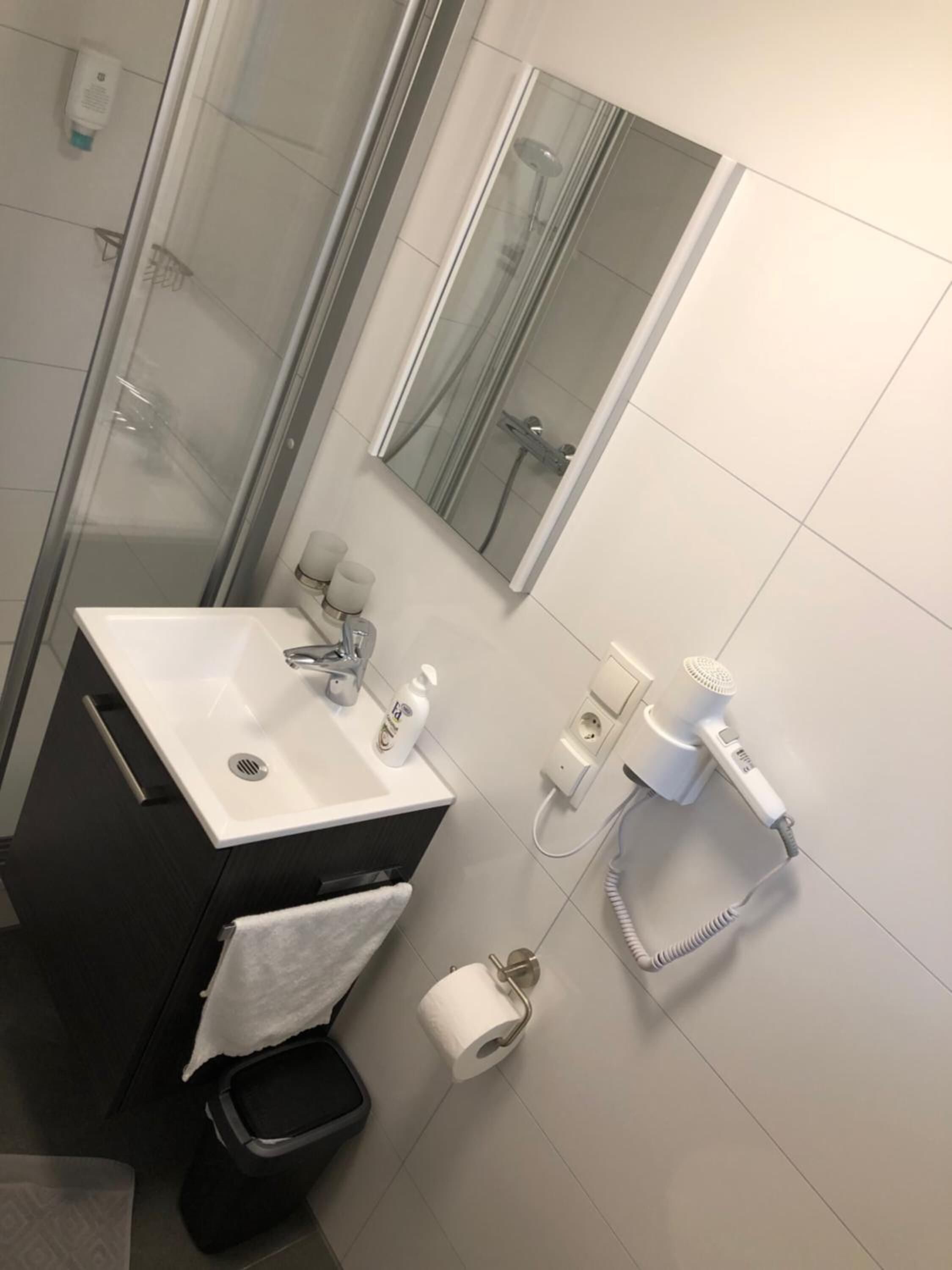A modern bathroom with a glass-enclosed shower, a white sink with a mirror above it, and a hairdryer on a wall socket.