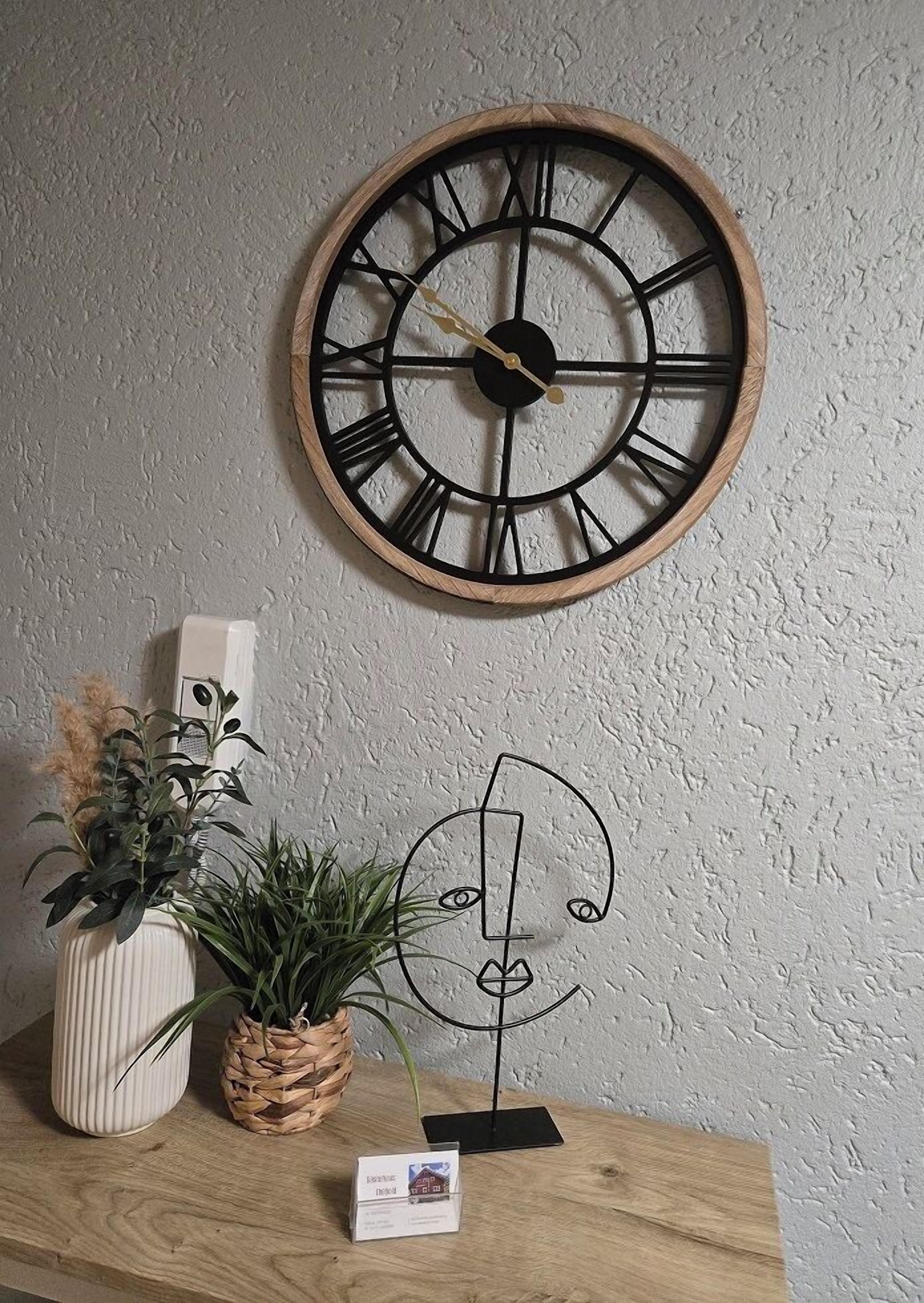 Une horloge murale en bois avec des chiffres romains, une petite plante et une œuvre d’art minimaliste, le tout posé sur une étagère en bois, devant un mur à la texture particulière.