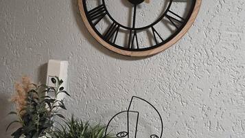 Une horloge murale en bois avec des chiffres romains, une petite plante et une œuvre d’art minimaliste, le tout posé sur une étagère en bois, devant un mur à la texture particulière.