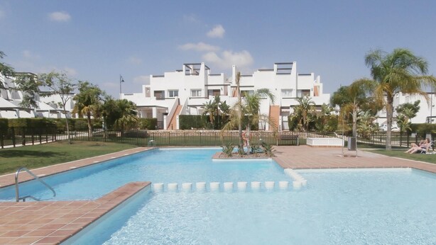 Una piscina con solárium de baldosas, rodeada de palmeras y un edificio blanco con balcones al fondo.